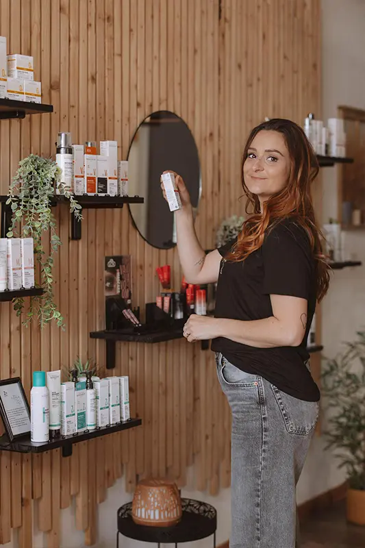 Institut-PourquoiPas_Bronzage_Naturel_BOTAN-01 Une femme en tenue décontractée sourit en choisissant un produit de soin sur une étagère. La présentation moderne présente divers produits et des plantes vertes, créant une atmosphère chaleureuse.