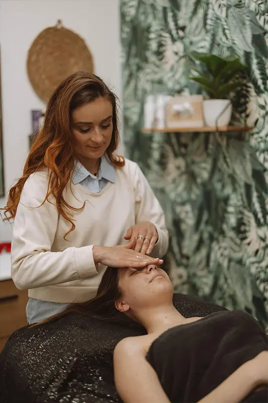 Institut-PourquoiPas_Expertise_soin_visage_Novexpert-01 Une femme reçoit un massage du visage apaisant dans un spa, où une esthéticienne lui exerce de légères pressions sur le front. La pièce est décorée dans un style botanique apaisant.