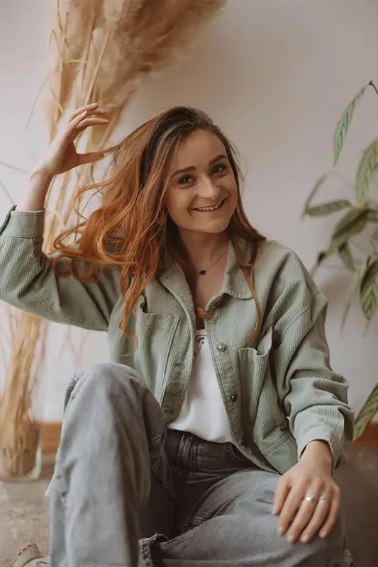 Insitut Pourquoi Pas A Propos Maeva Arnouts-2 Une femme souriante, vêtue d'une veste verte et d'un jean, est assise tranquillement à l'intérieur. Elle relève joyeusement ses cheveux, entourée d'herbes de la pampa et d'une plante. Ambiance chaleureuse et joyeuse.