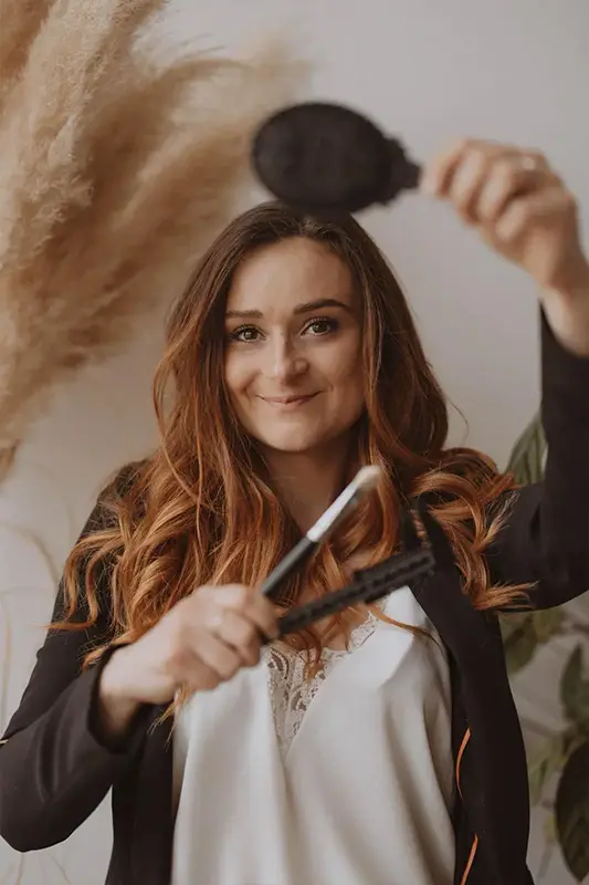 Insitut Pourquoi Pas A Propos Maeva Arnouts-3 Une femme souriante aux cheveux longs et ondulés tient un pinceau à maquillage et une brosse à cheveux. Elle se trouve dans un décor chaleureux, avec de l'herbe de la pampa en arrière-plan, créant une ambiance chaleureuse et accueillante.